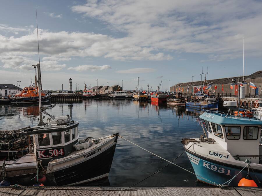 A harbor with boats docked at the pier, a lighthouse in the background, and buildings along the waterfront.
