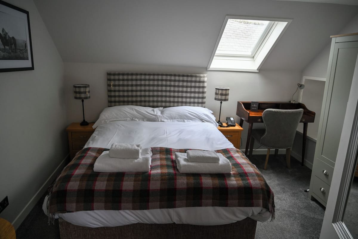 A neatly made bed with a plaid blanket and two folded towels, a desk with a chair, a lamp, and a wardrobe in a hotel room.