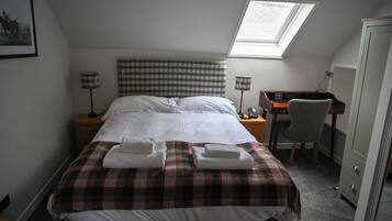 Una cama bien hecha con una manta escocesa y dos toallas dobladas, un escritorio con una silla, una lámpara y un armario en una habitación de hotel.