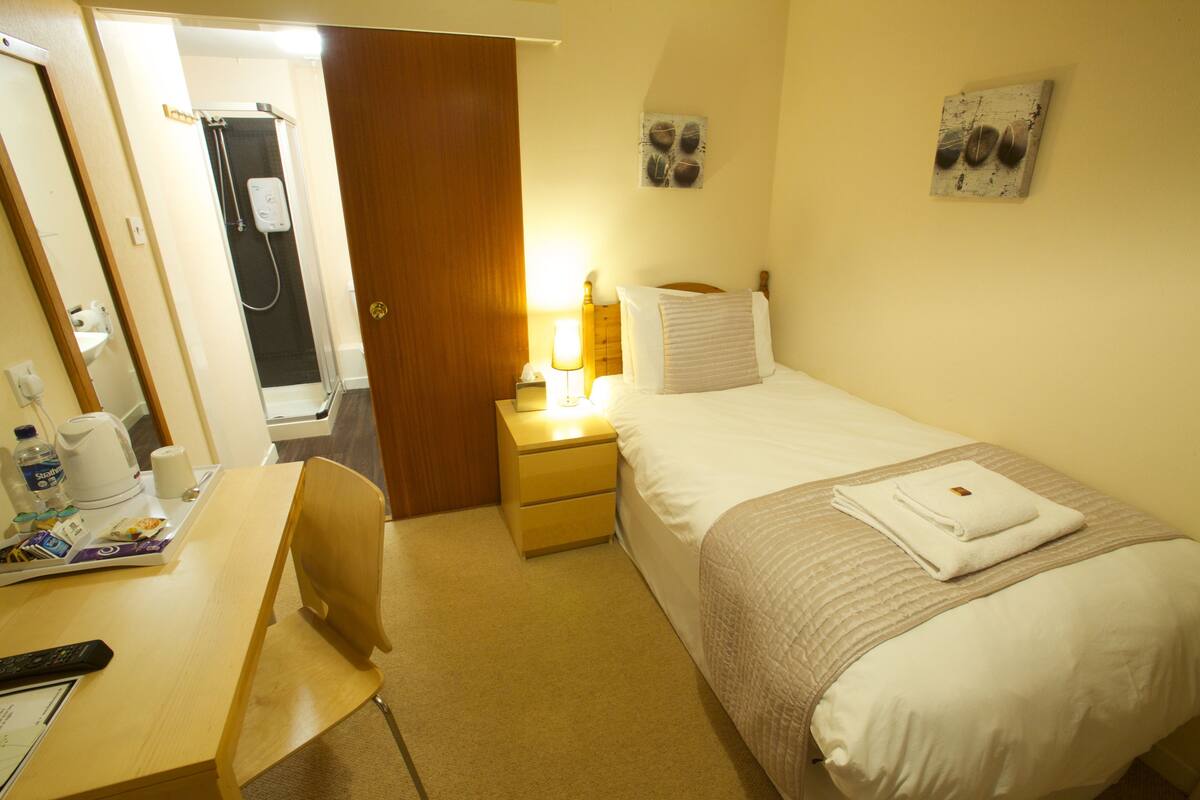 A hotel room with a bed, bedside table, chair, and a shower area.