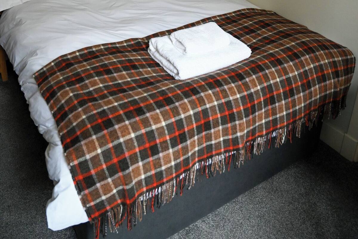 A neatly made bed with a plaid blanket and a folded white towel, framed black-and-white photographs on the wall, and a dark headboard.