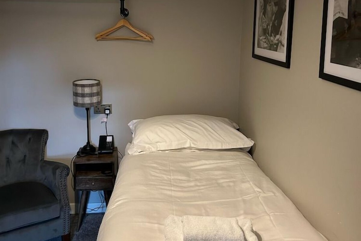 Un dormitorio con cama, una silla, una mesita de noche con lámpara y un perchero.