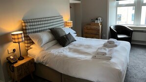 Ein ordentlich bezogenes Bett mit weißen Leinen und einem karierten Kopfteil, eine Nachttischlampe, ein Holznachttisch mit einem Telefon und ein Stuhl am Fenster.