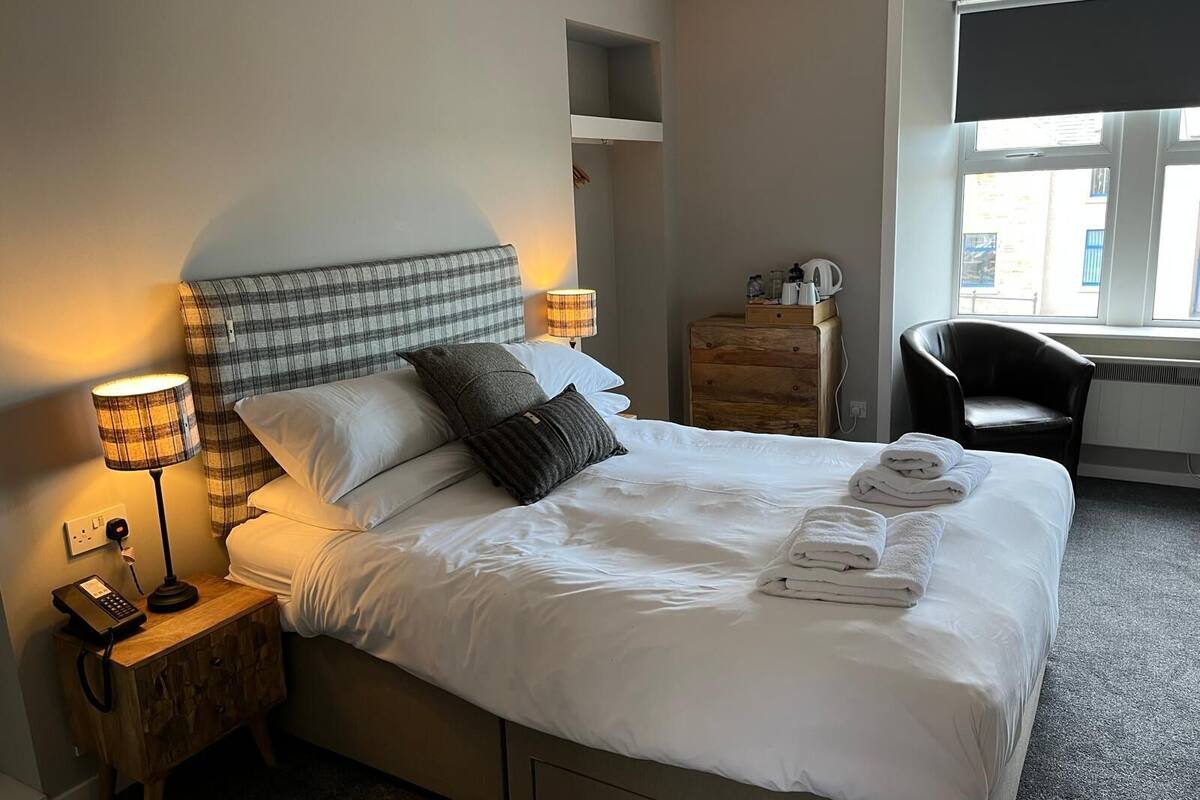 A neatly made bed with white linens and a plaid headboard, a bedside lamp, a wooden nightstand with a phone, and a chair by a window.