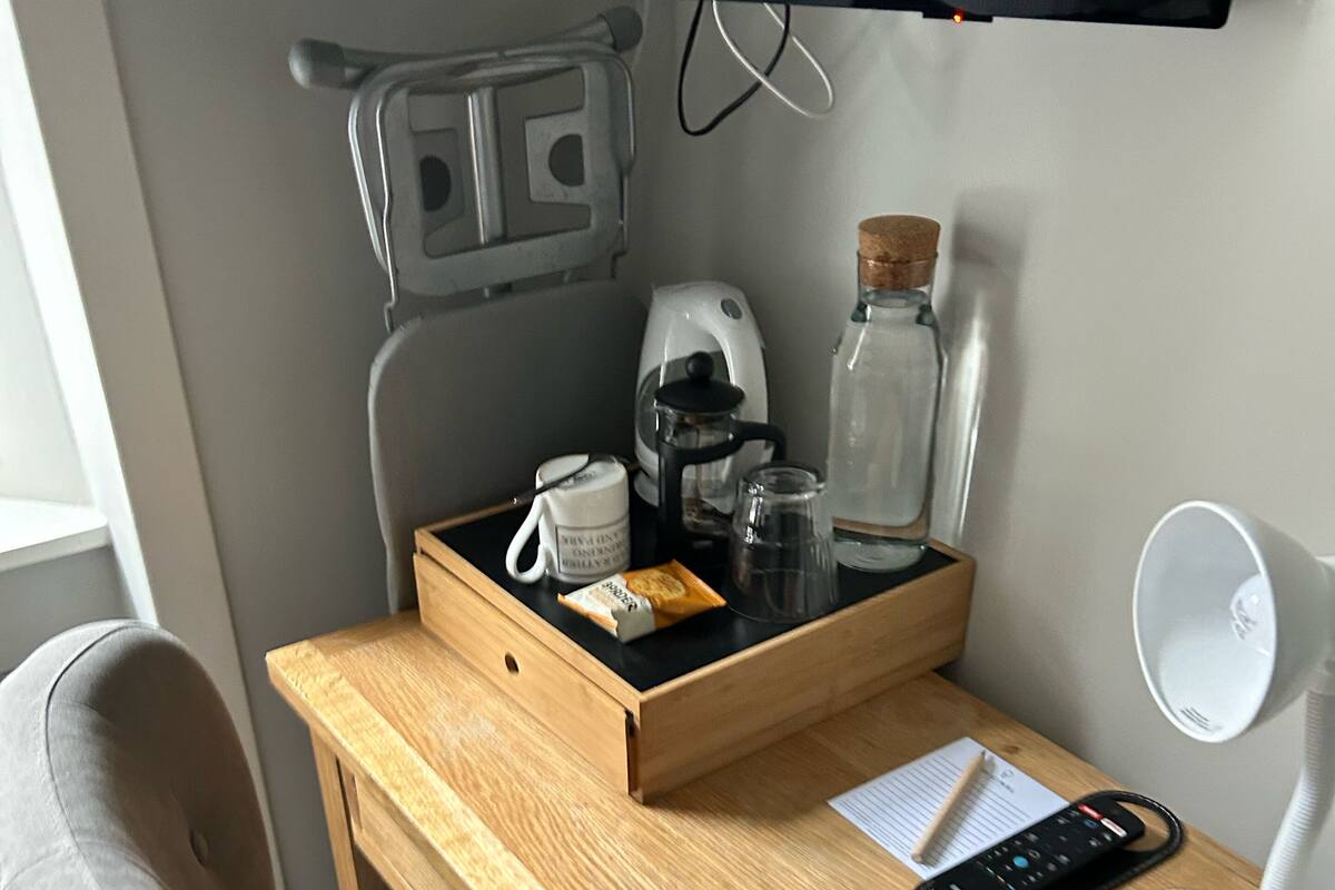 A wooden desk with a computer monitor, a coffee maker, a kettle, a glass jar, a mug, a remote control, and a desk lamp.