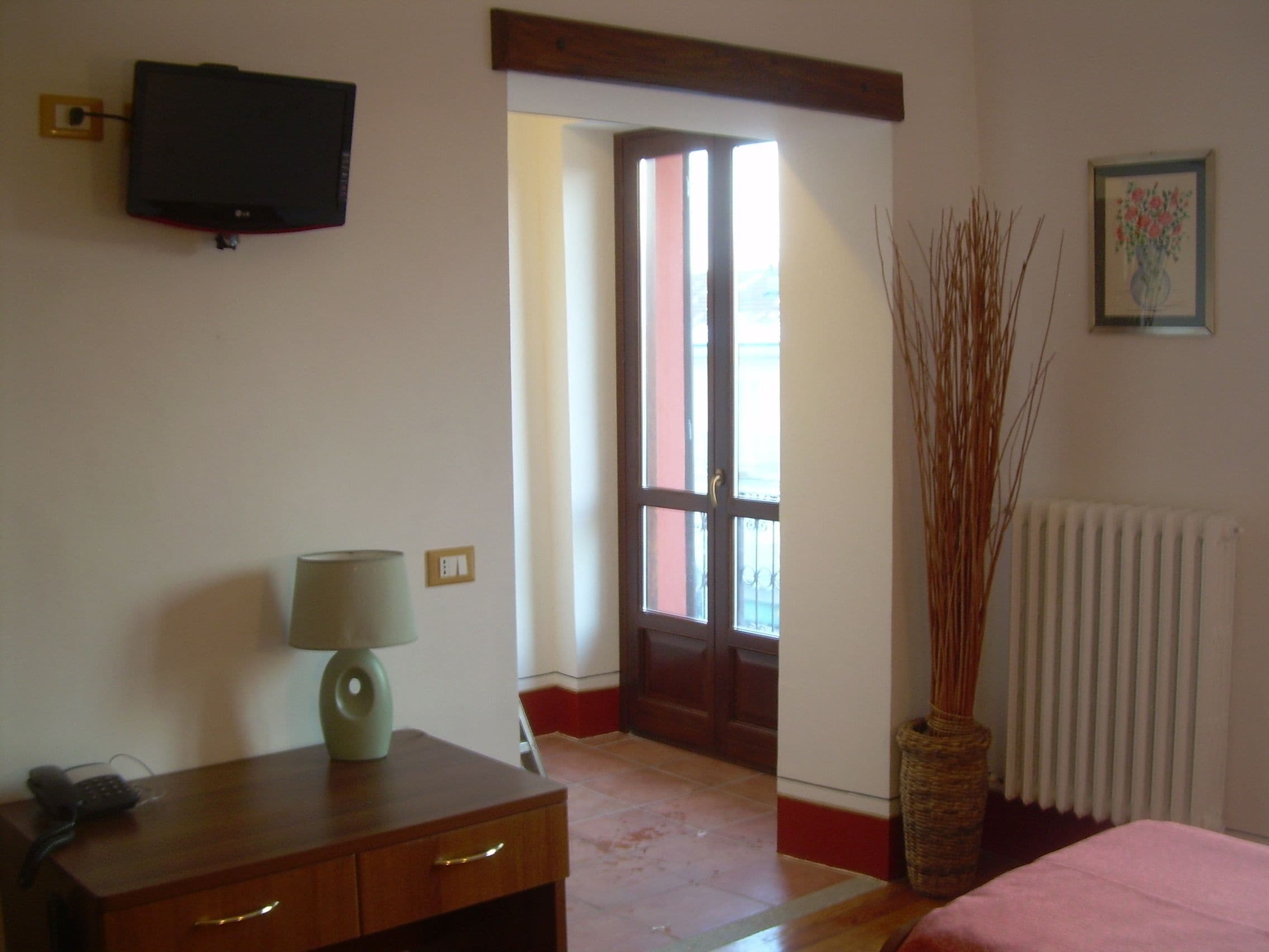 Una habitación con un escritorio de madera, una lámpara, un televisor en la pared, una puerta que da al exterior, un radiador y un jarrón con plantas altas.