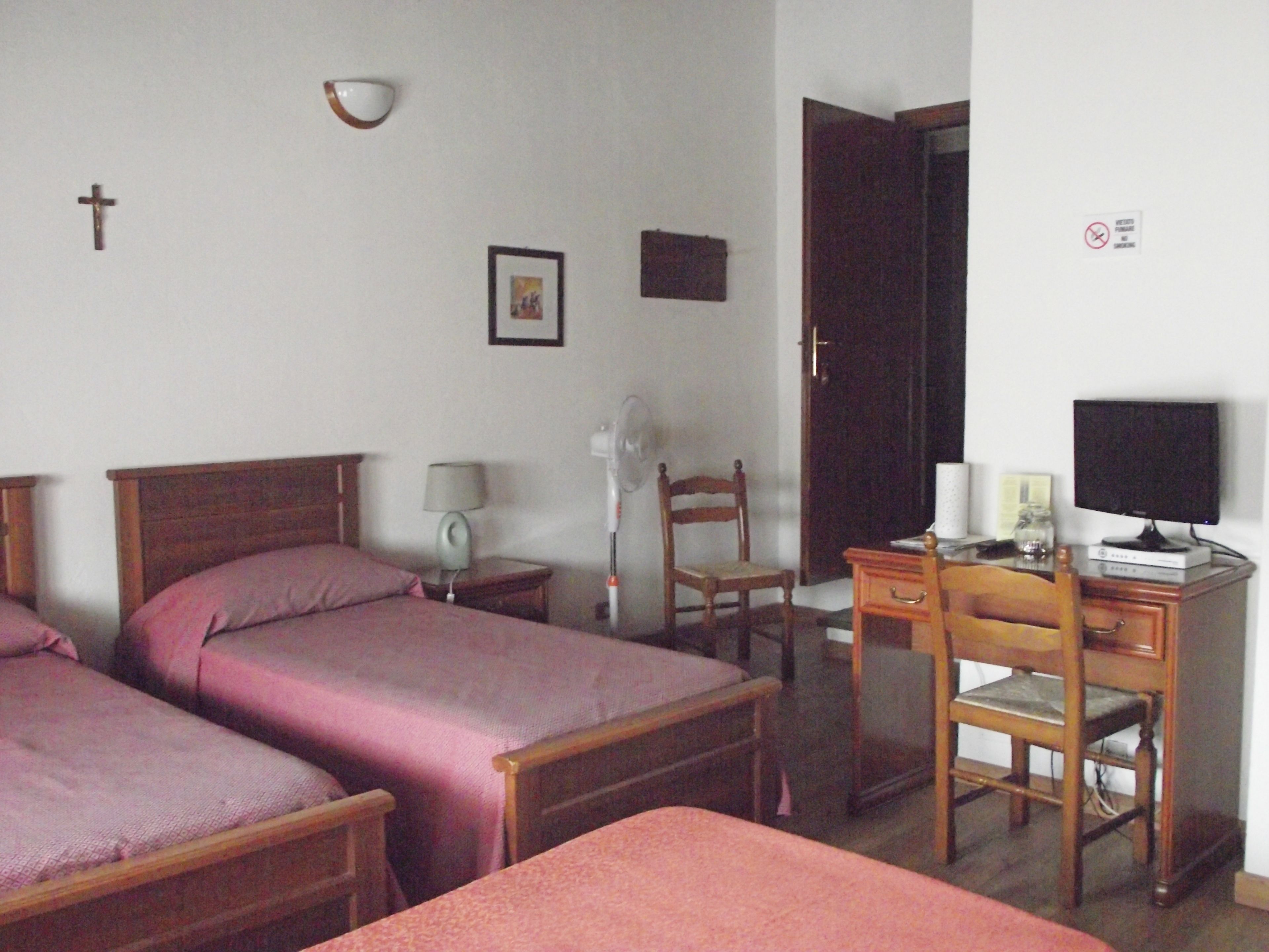 Quarto com duas camas de madeira, uma mesa com cadeira, luminária de mesa, ventoinha na parede e uma televisão.
