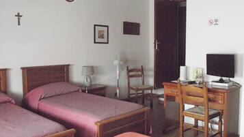 Quarto com duas camas de madeira, uma mesa com cadeira, luminária de mesa, ventoinha na parede e uma televisão.