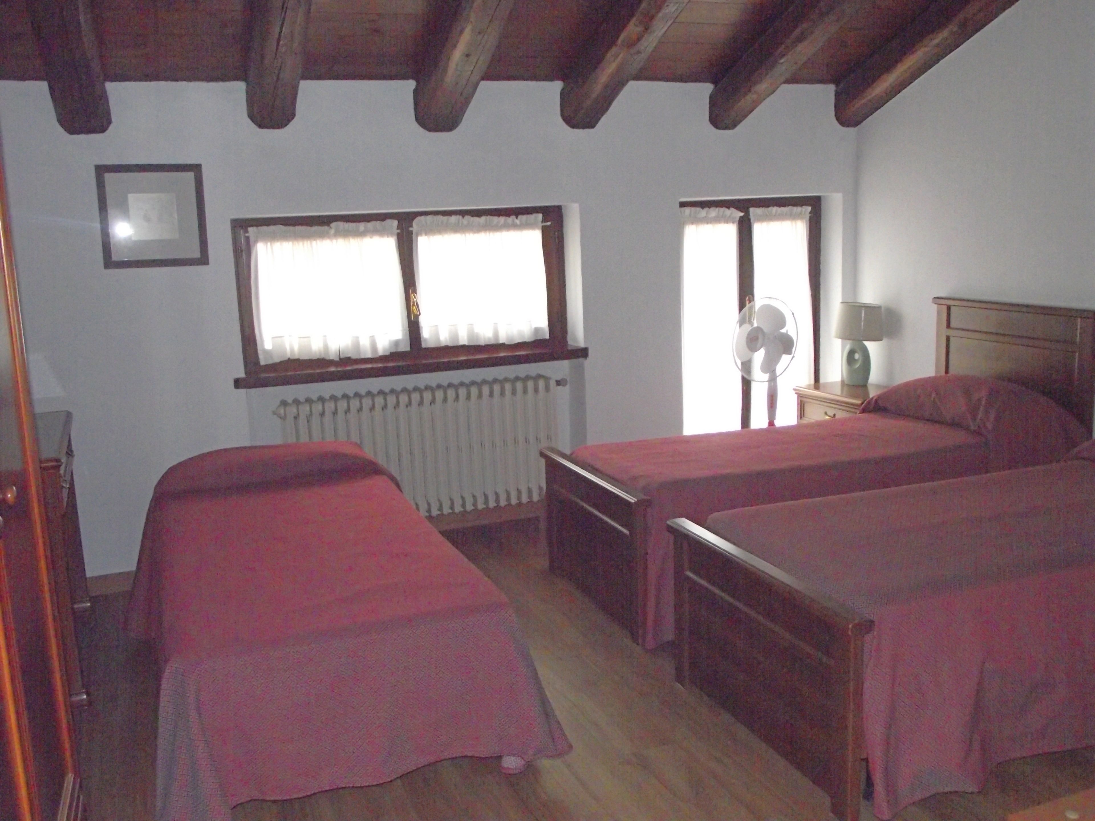 Una habitación con dos camas individuales, un radiador, una ventana con cortinas, un ventilador y una mesita de noche con lámpara.