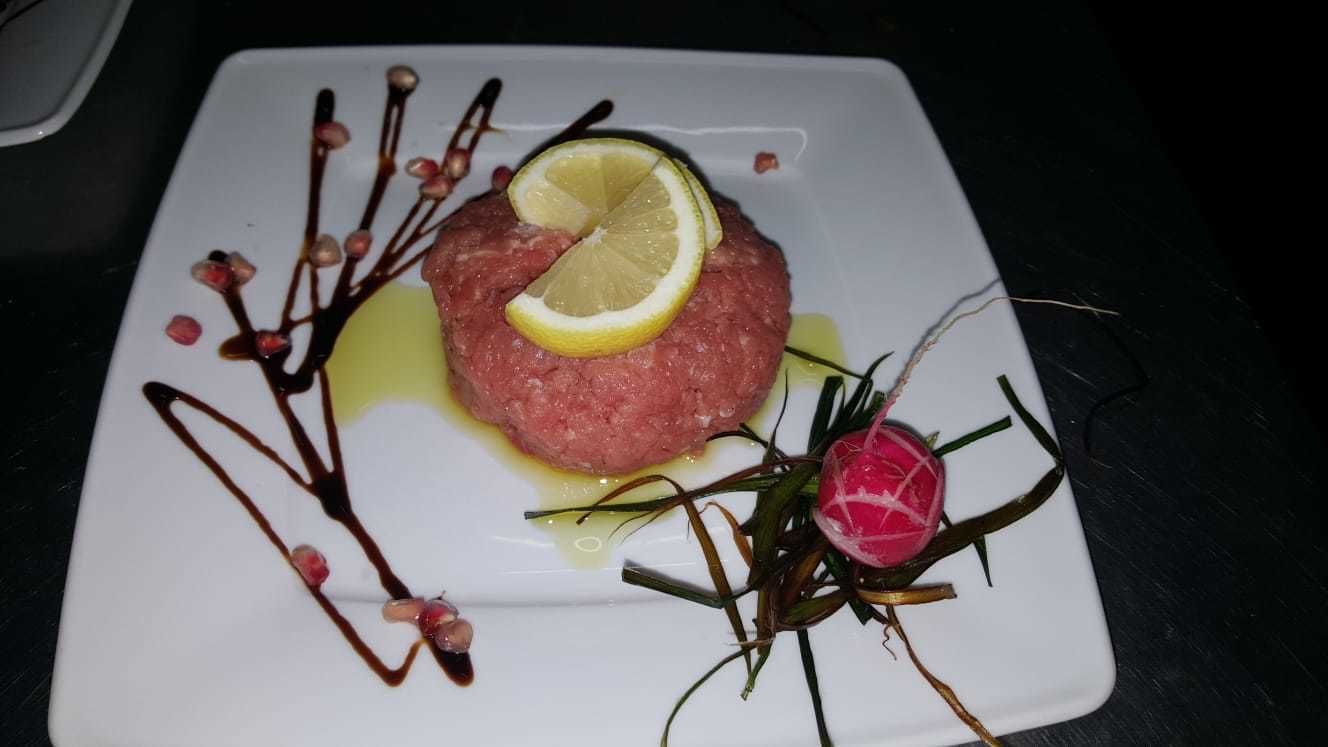 Un plato cuadrado con un guiso de carne cruda, una rodaja de limón y elementos decorativos.