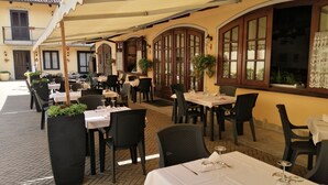 Un'area pranzo all'aperto con tavoli e sedie, piante in vaso e un edificio con finestre ad arco e tetto in tegole.