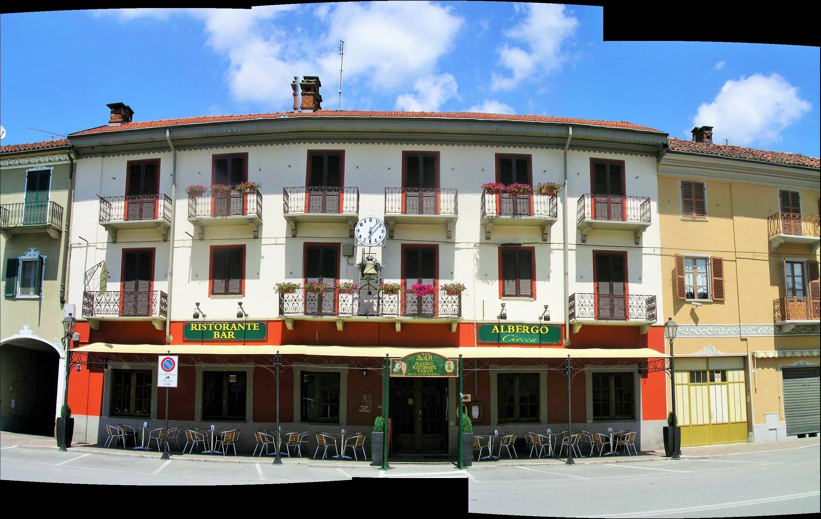 Un edificio de varios pisos con un bar y restaurante en la planta baja, balcones con jardineras florales y un cielo azul despejado.