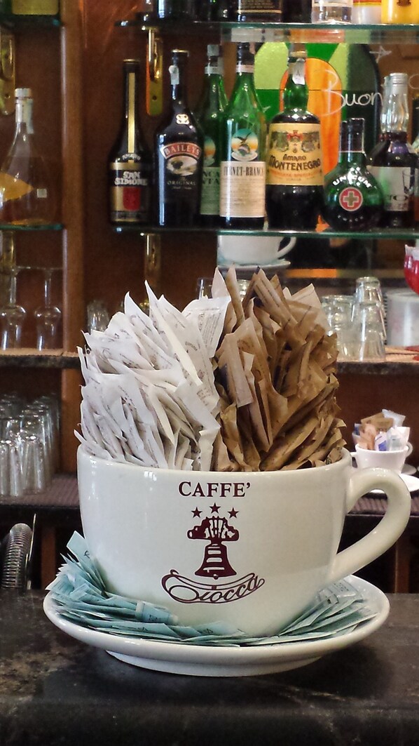 A white coffee cup with 'CAFFE' and a bell logo, filled with coffee capsules and a saucer. - Albergo Ciocca (Castelnuovo don Bosco)