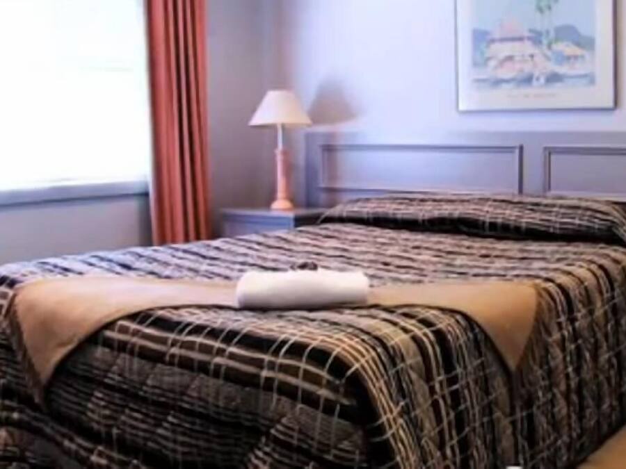 Una cama bien hecha con una colcha escocesa, una lámpara de noche y un cuadro en la pared.