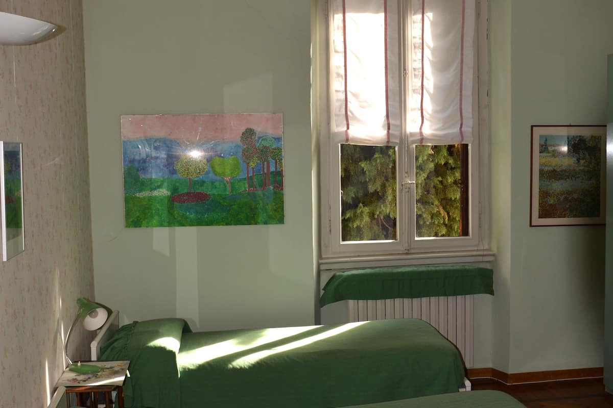 A bedroom with two single beds, a window with curtains, a desk with a lamp, and two paintings on the wall.