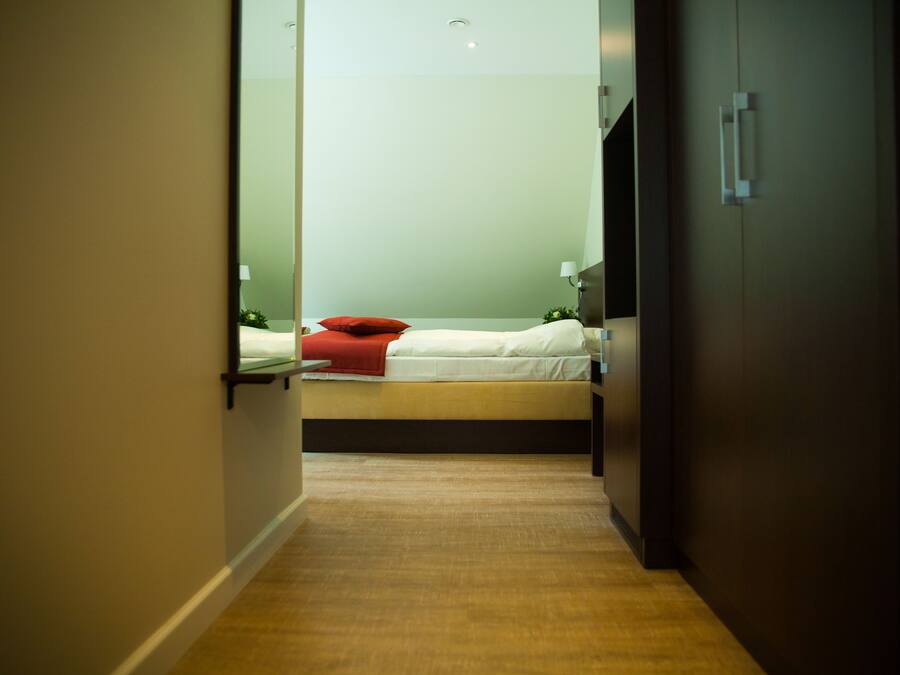 A narrow hallway leading to a bed with a red pillow, a bedside table with a lamp, and a wall-mounted light fixture.