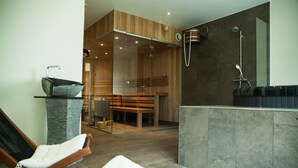 A modern bathroom with a glass-enclosed shower, a wooden bench, and a seating area with a cushioned chair. - Hotel BramslevGaard (Hobro)