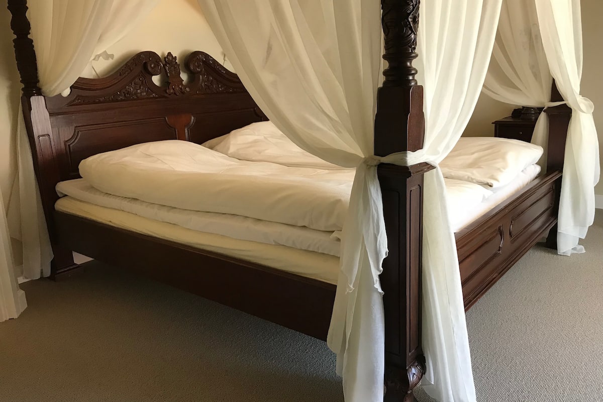 A four-poster canopy bed with white linens, wooden headboard, and a nightstand with a lamp.