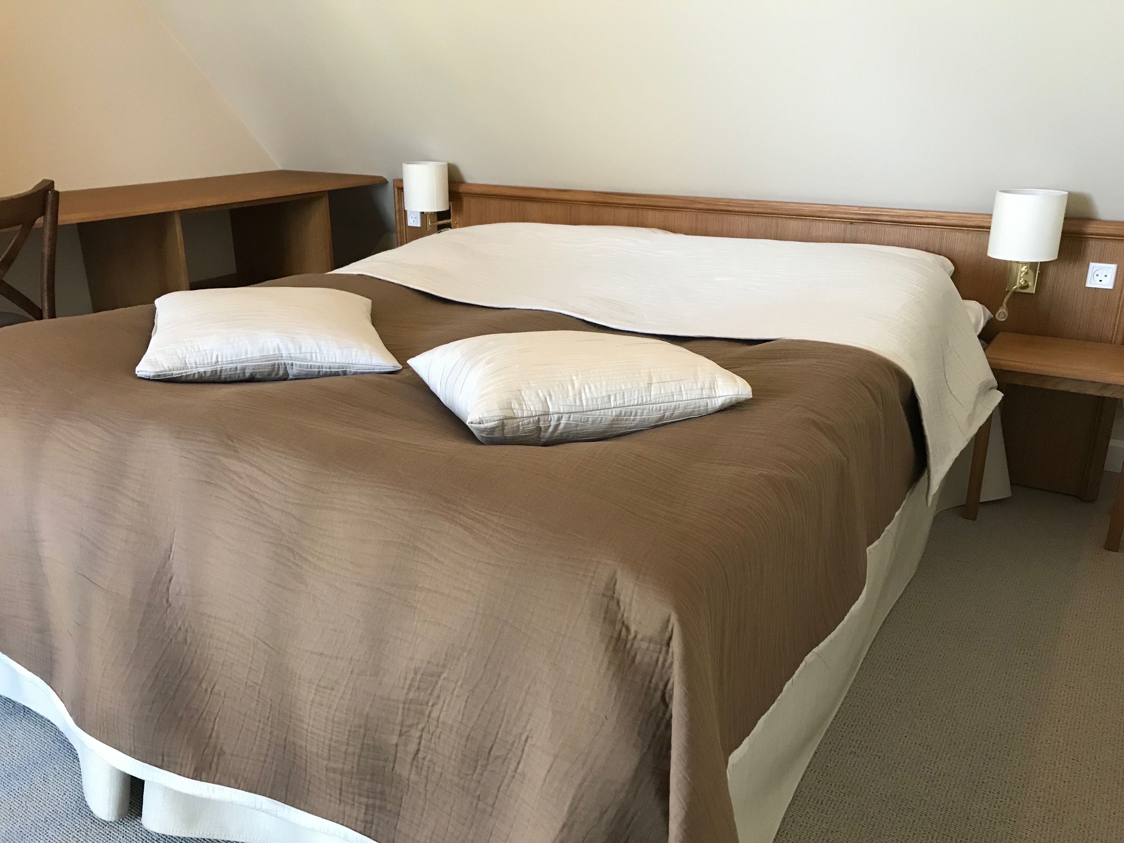 Un lit bien fait, recouvert d’une couverture marron et agrémenté de deux oreillers, une table de chevet en bois avec une lampe, et une chaise en bois.