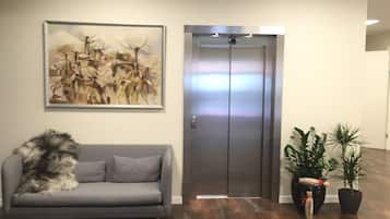 A lobby with a grey sofa, a painting, an elevator, and potted plants.