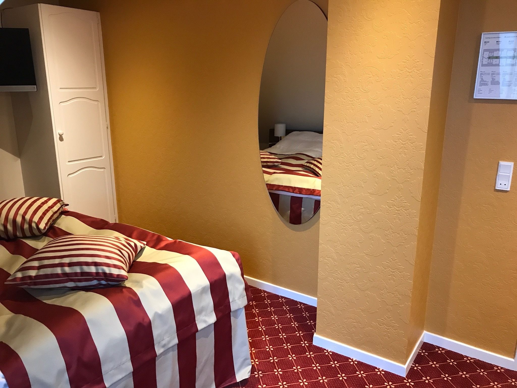 A room with a bed featuring a striped bedspread, a white wardrobe, a mirror on the wall, and a patterned floor.