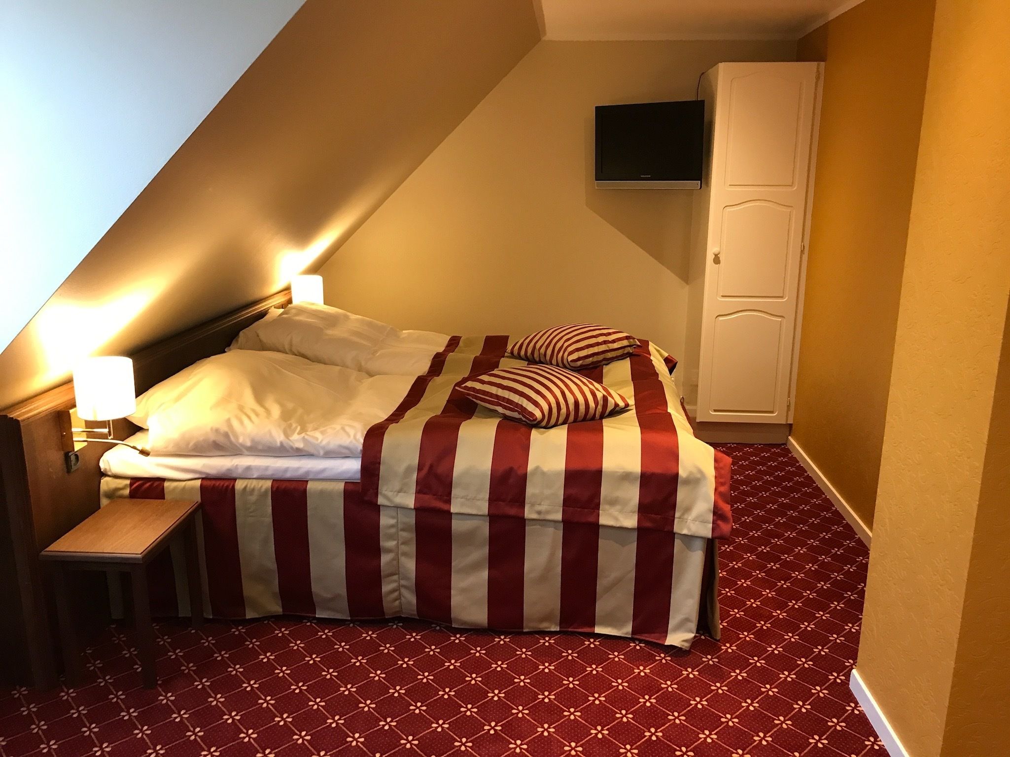 A hotel room with a bed, a nightstand, a lamp, a wardrobe, and a TV mounted on the wall.
