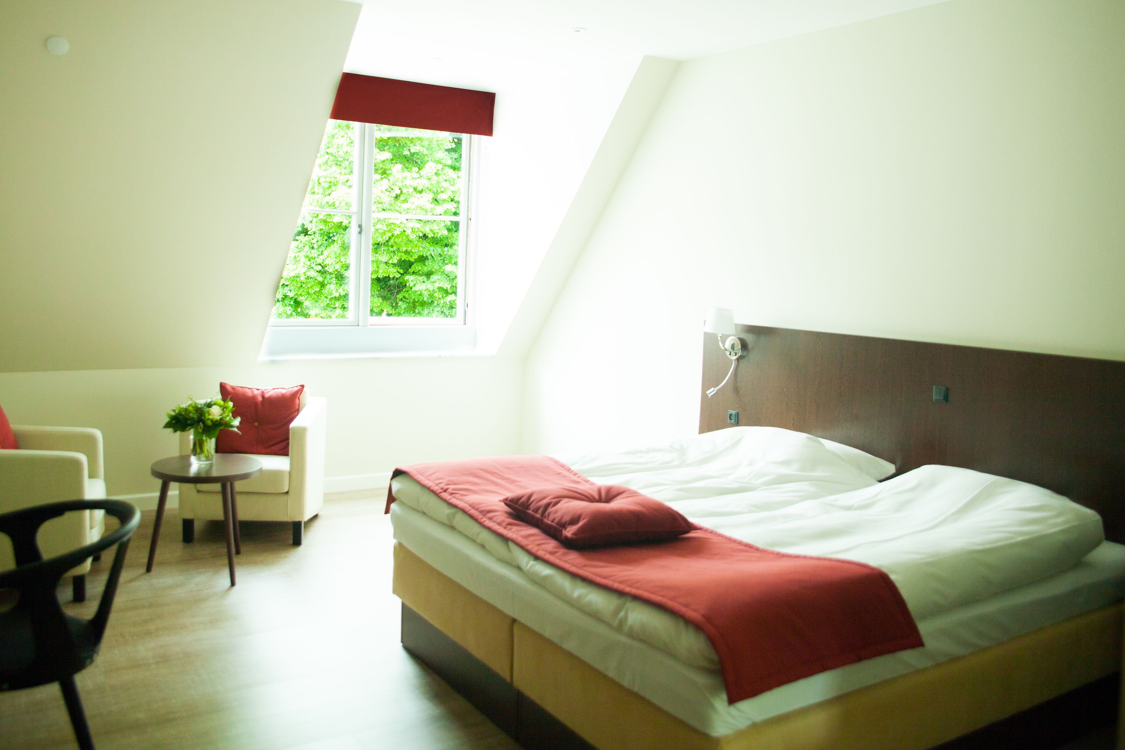 A bedroom with a bed, a chair, a small table with a plant, and a window with a view of greenery.