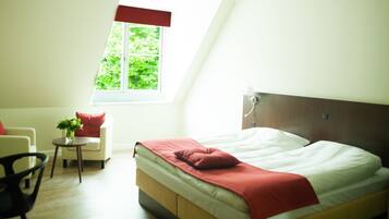 A bedroom with a bed, a chair, a small table with a plant, and a window with a view of greenery.