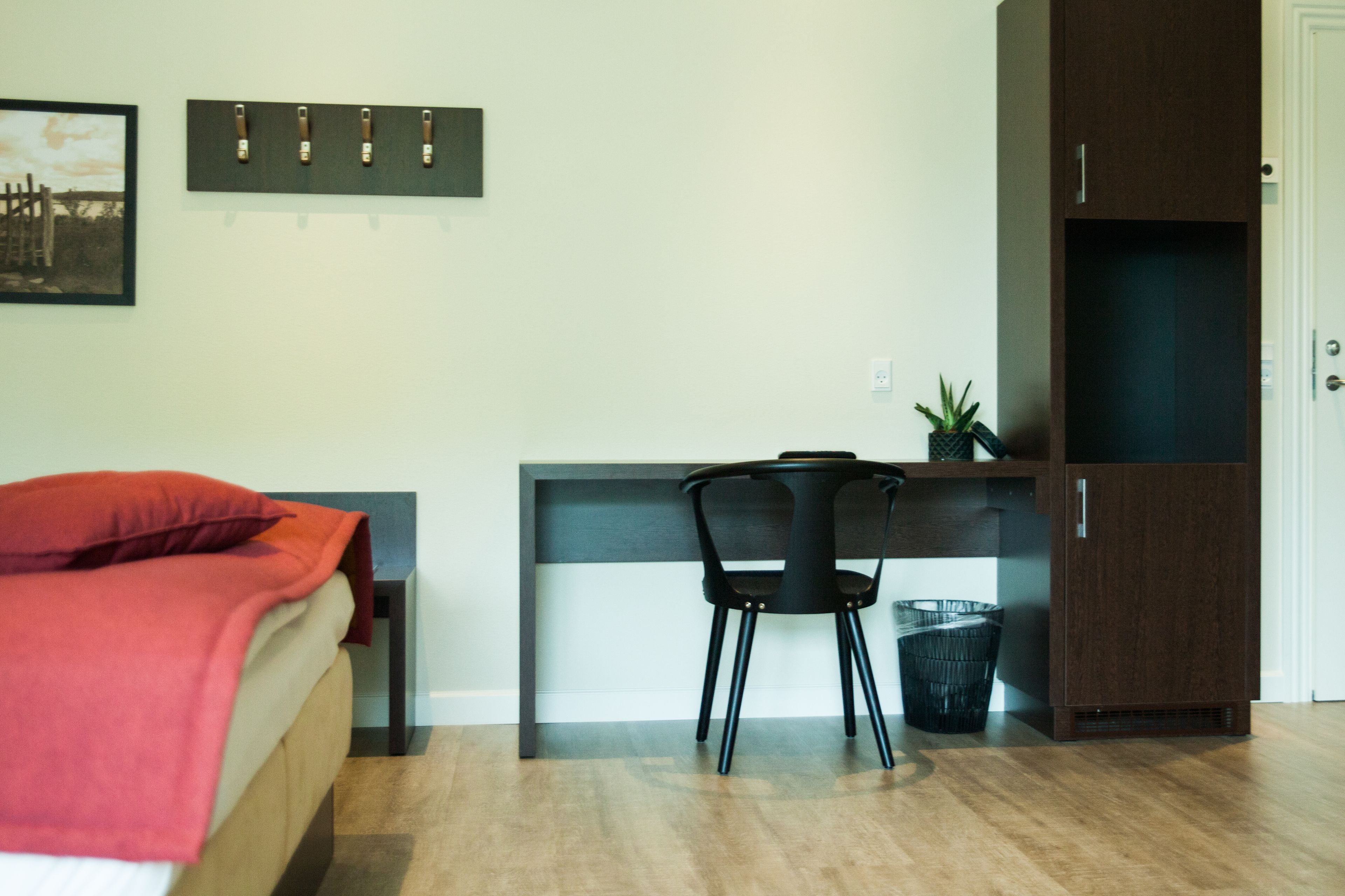 A bedroom with a bed, a desk, a chair, a wardrobe, and a small plant.