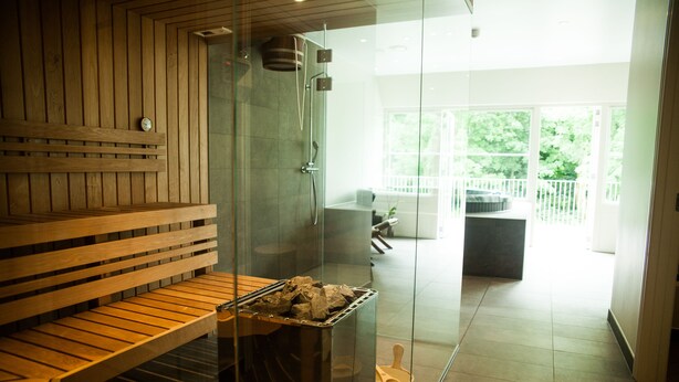 A sauna room with a wooden bench, a glass shower enclosure, and a view of a balcony with outdoor furniture. - Hotel BramslevGaard (Hobro)