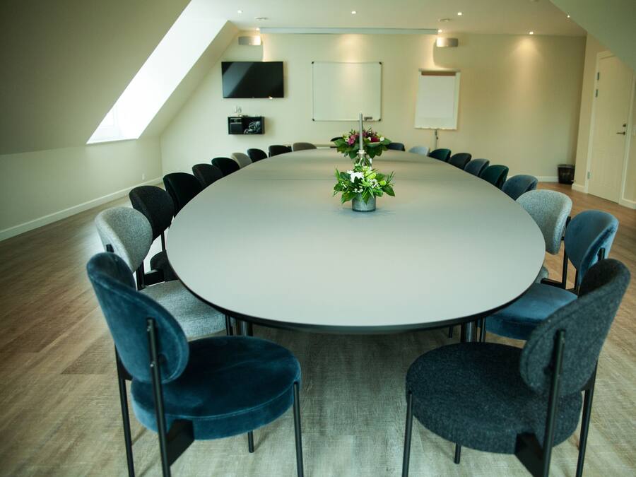 Ein Konferenzraum mit rundem Tisch, Stühlen, einer Blumenvase und einem Fernseher an der Wand.