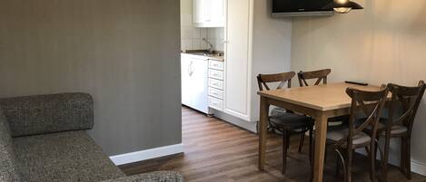 A compact living space with a sofa, dining table, and chairs, a kitchen area with cabinets and a TV, and a wooden floor.