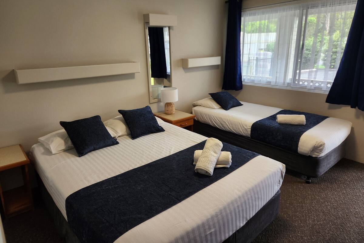 A hotel room with two beds, a bedside table with a lamp, a mirror, and a window with curtains.