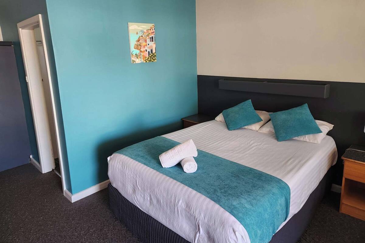 A hotel room with a bed, a nightstand, a painting on the wall, and a door to the left.