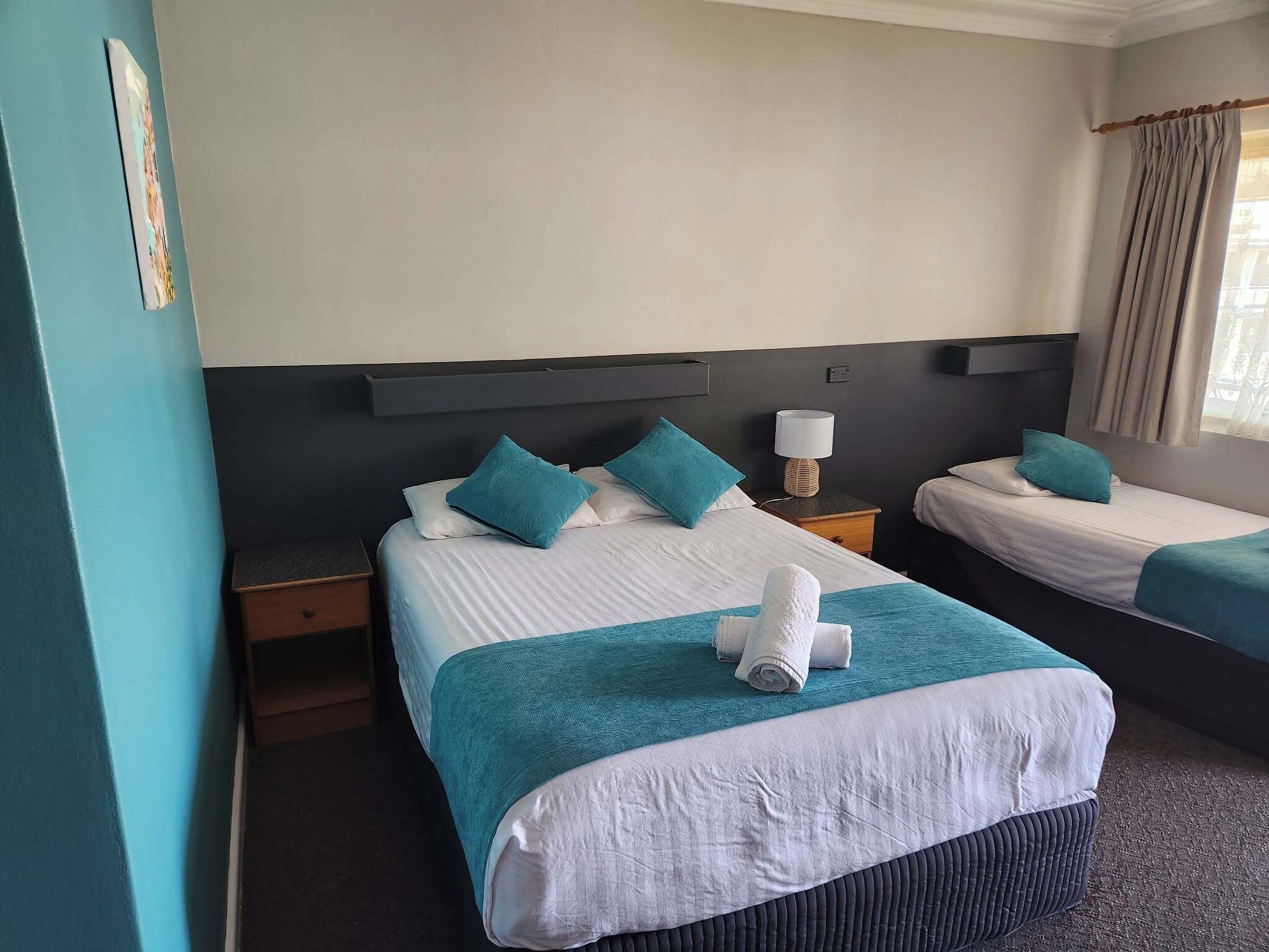 A hotel room with two beds, each with a teal bedspread and white pillows. There is a nightstand with a lamp between the beds.