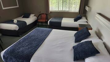 A hotel room with two single beds and one double bed, all with blue bedding. There is a chair and a small table in the room.