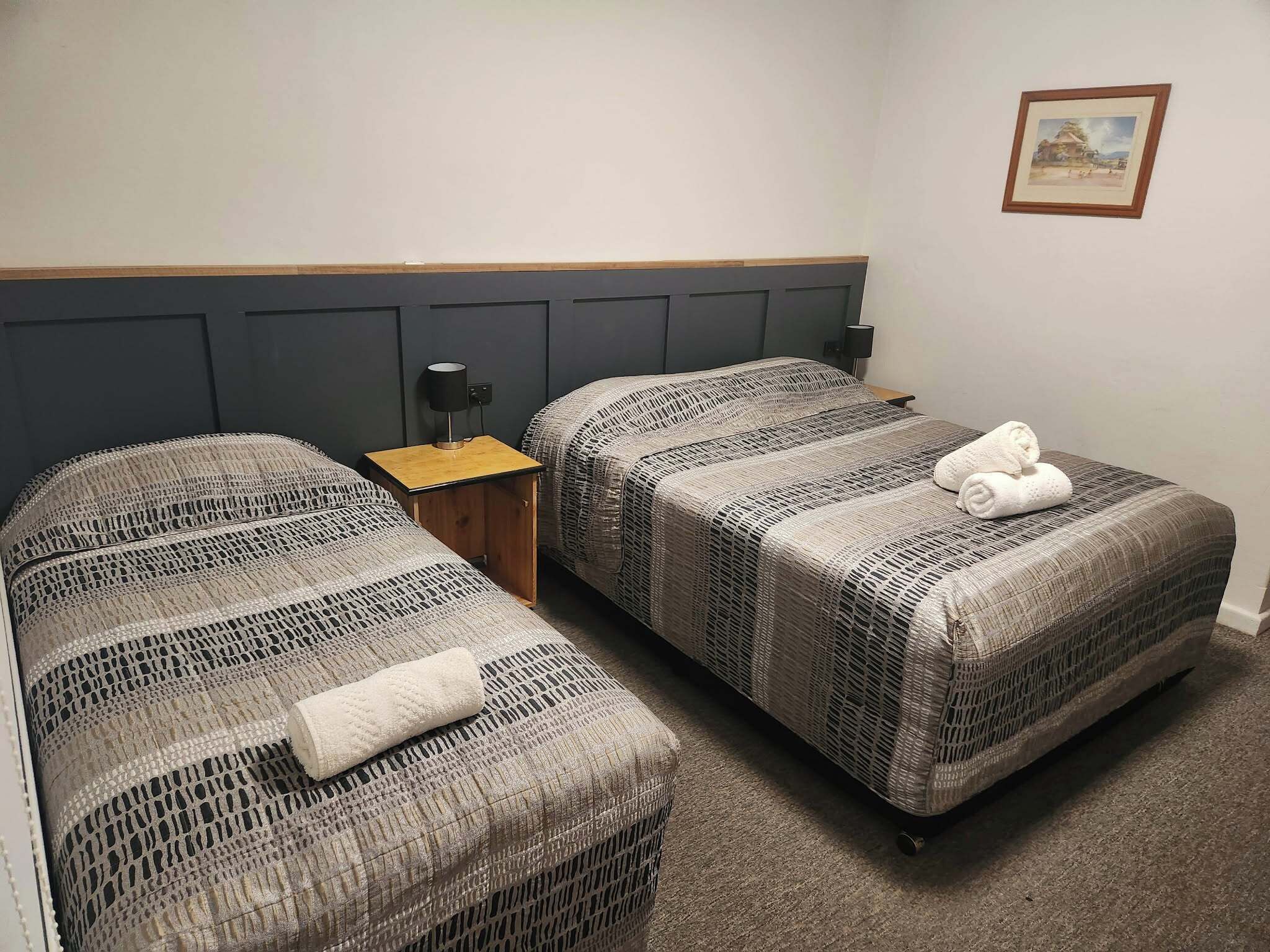 Una habitación doble con dos camas individuales, una pequeña mesita de noche con lámpara, y un cuadro en la pared.