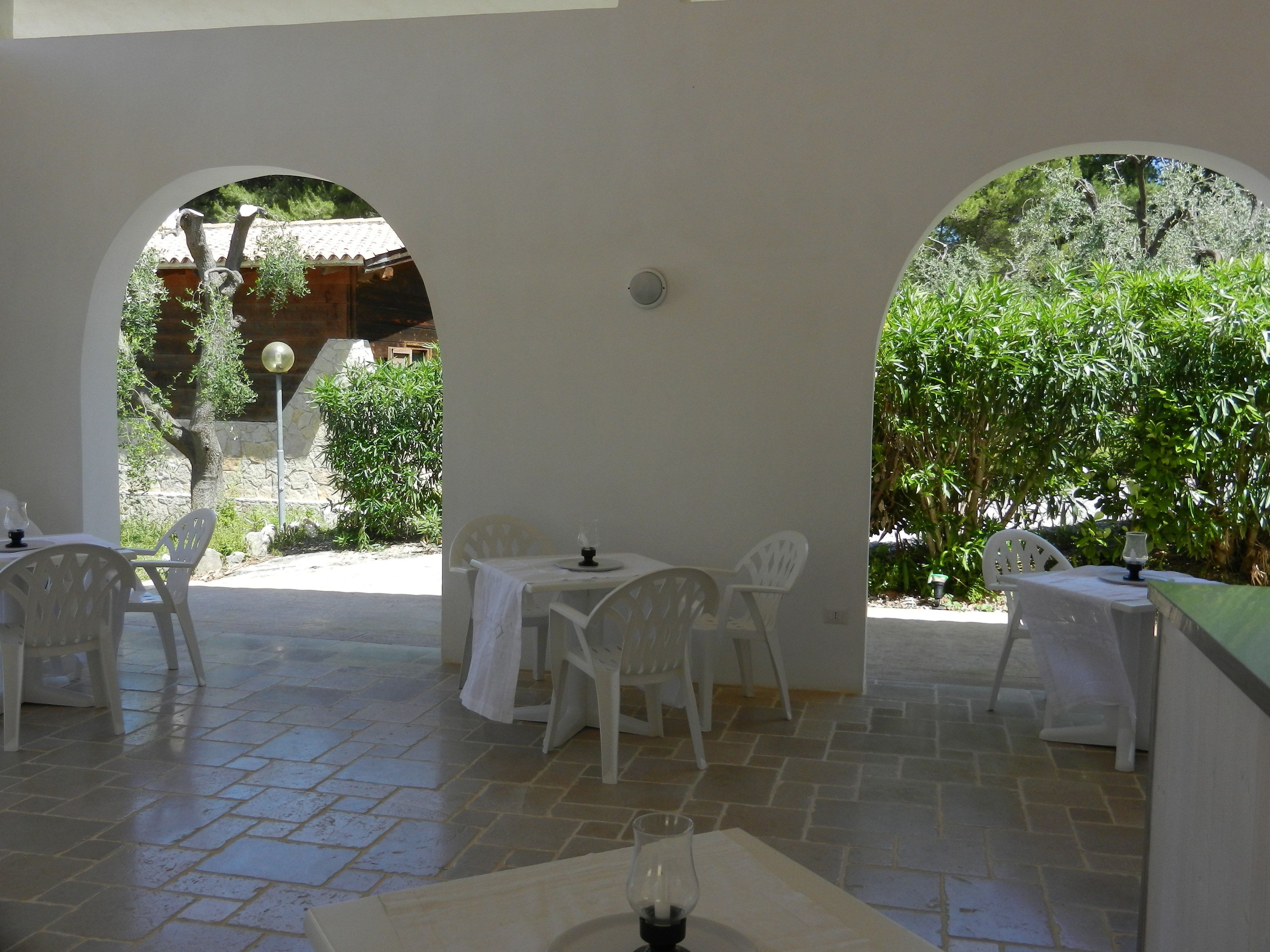 Un patio al aire libre con mesas y sillas blancas, suelo de piedra y follaje en el fondo.