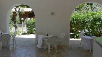 Un patio al aire libre con mesas y sillas blancas, suelo de piedra y follaje en el fondo.