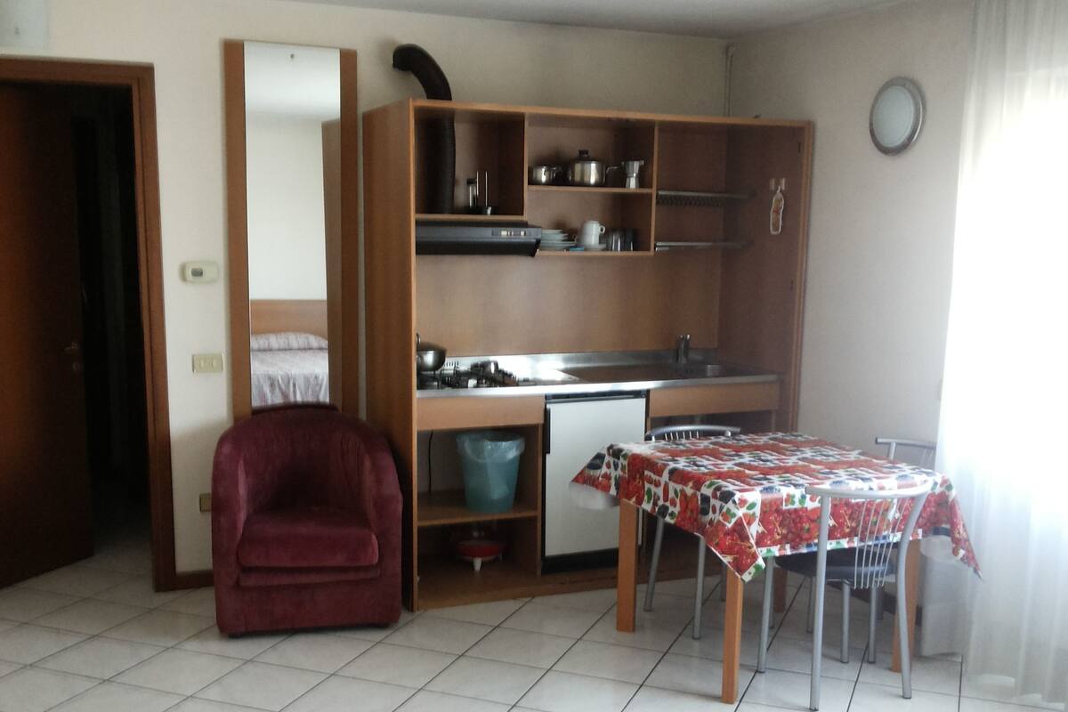 Una habitación compacta con kitchenette, una mesa de comedor con sillas, un sillón rojo y una cama.