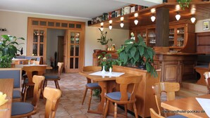 A wooden dining area with tables and chairs, a bar area with shelves of books and glasses, and a large potted plant. - Landhotel Pockau (Lengefeld)