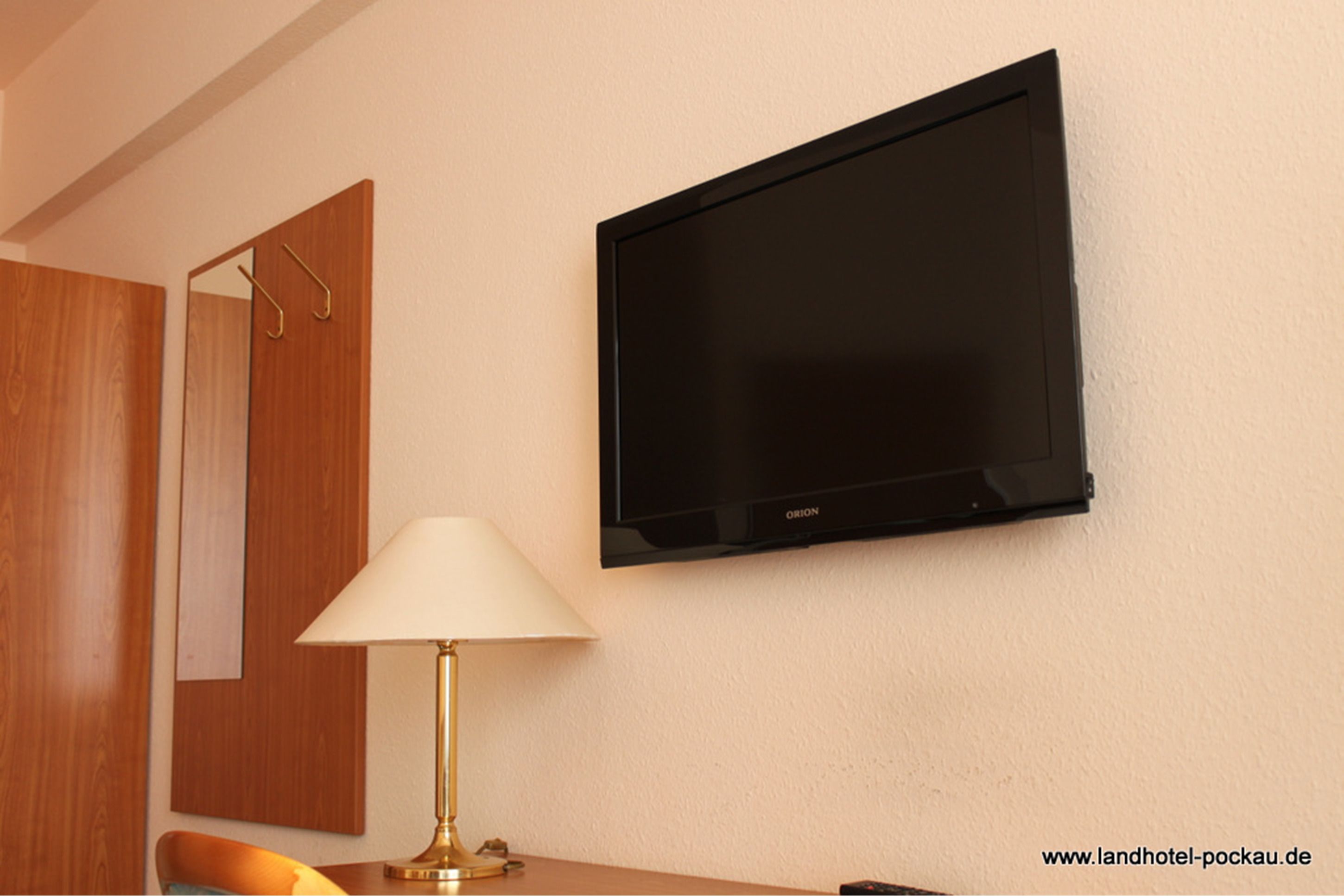A hotel room with a mounted flat-screen TV, a wooden wardrobe, a table with a lamp, and a chair.