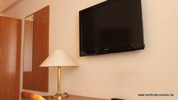 A hotel room with a mounted flat-screen TV, a wooden wardrobe, a table with a lamp, and a chair.
