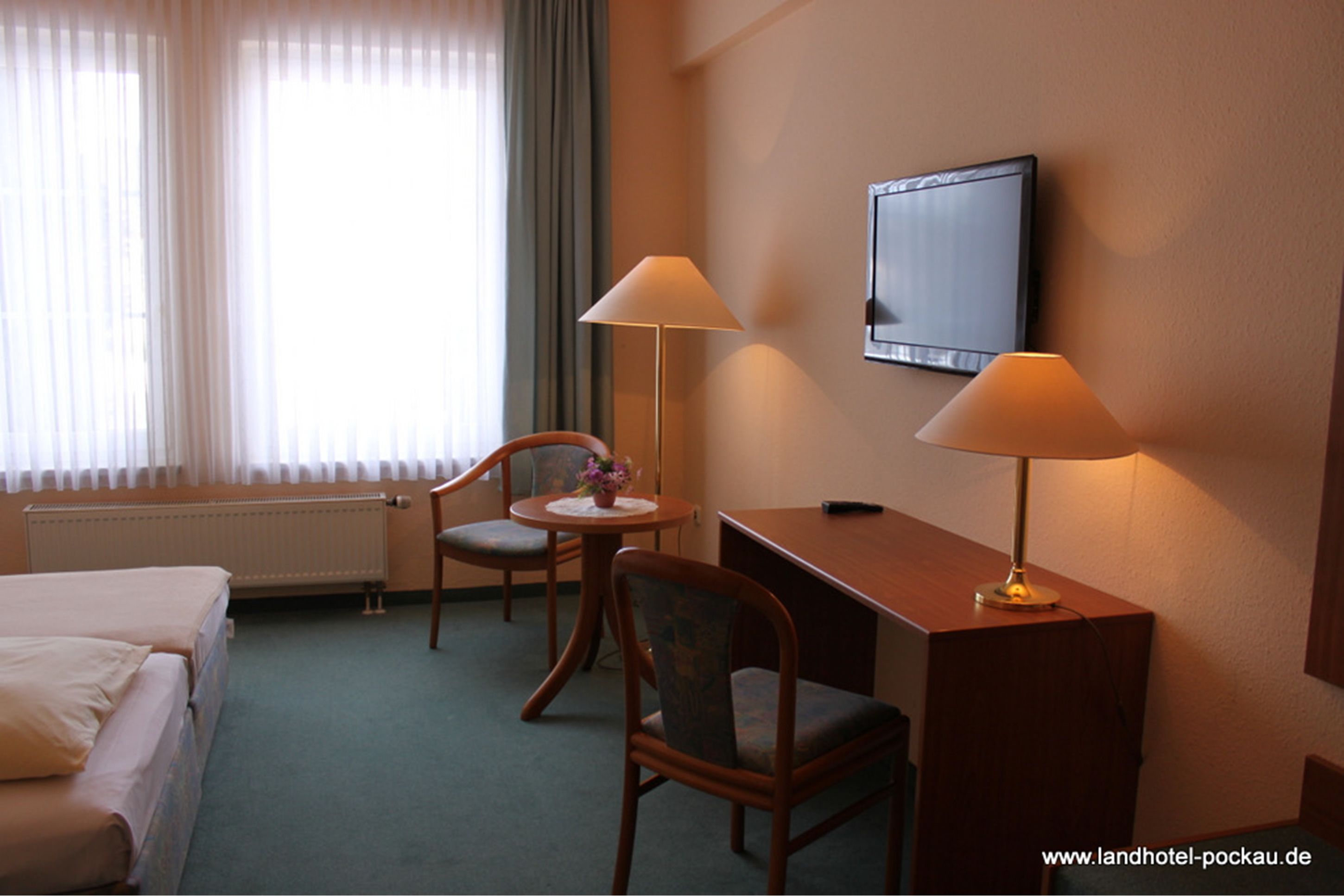 A hotel room with a bed, two chairs, a desk, a lamp, a TV, and a window with curtains.