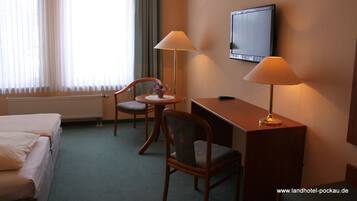 A hotel room with a bed, two chairs, a desk, a lamp, a TV, and a window with curtains.