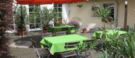 Un patio al aire libre con mesas y sillas, cubierto por un paraguas grande, rodeado de macetas con plantas y flores.