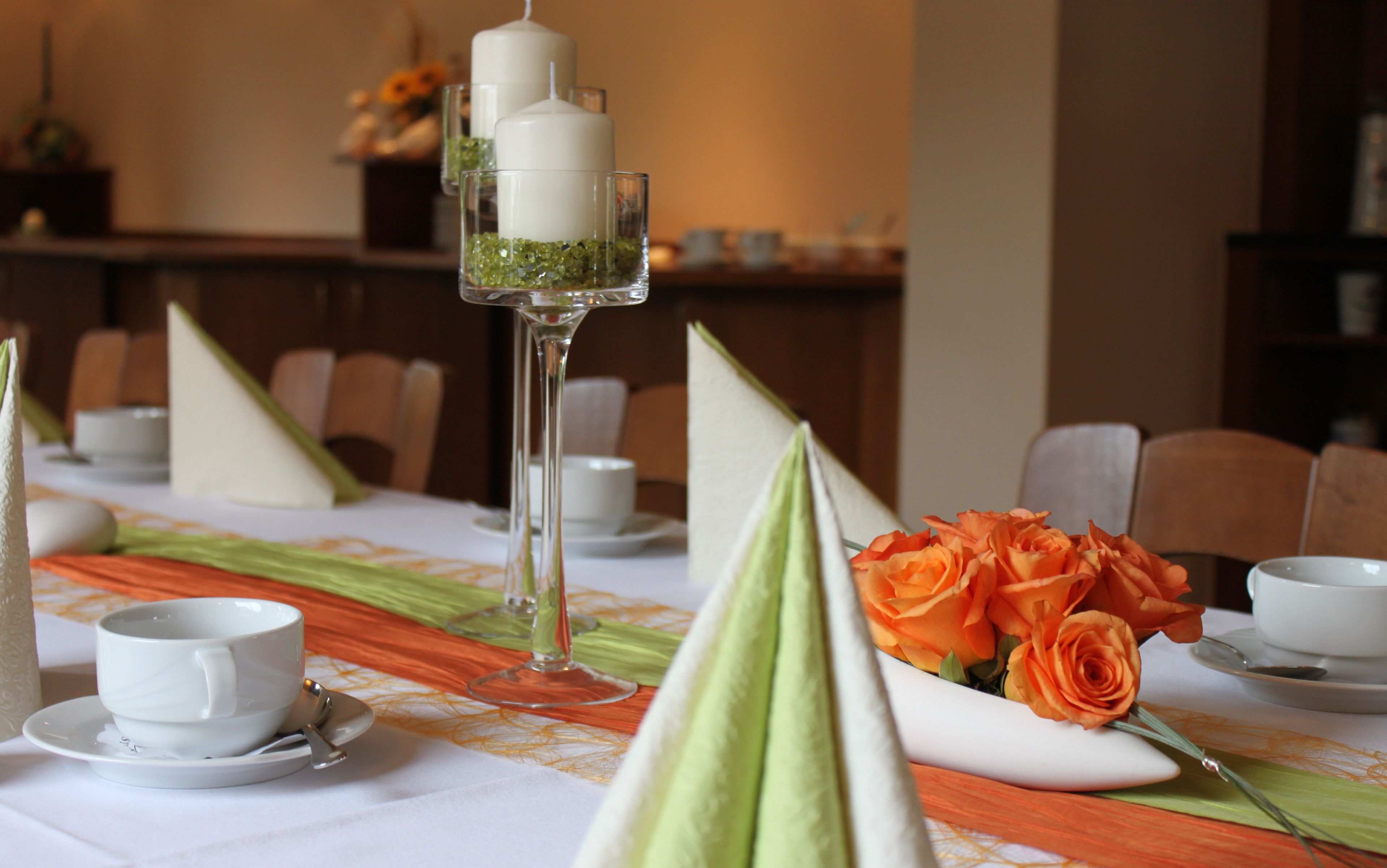 Una mesa de comedor dispuesta con tazas y platillos blancos, y un vaso con una guarnición verde. Hay rosas anaranjadas sobre un plato blanco.