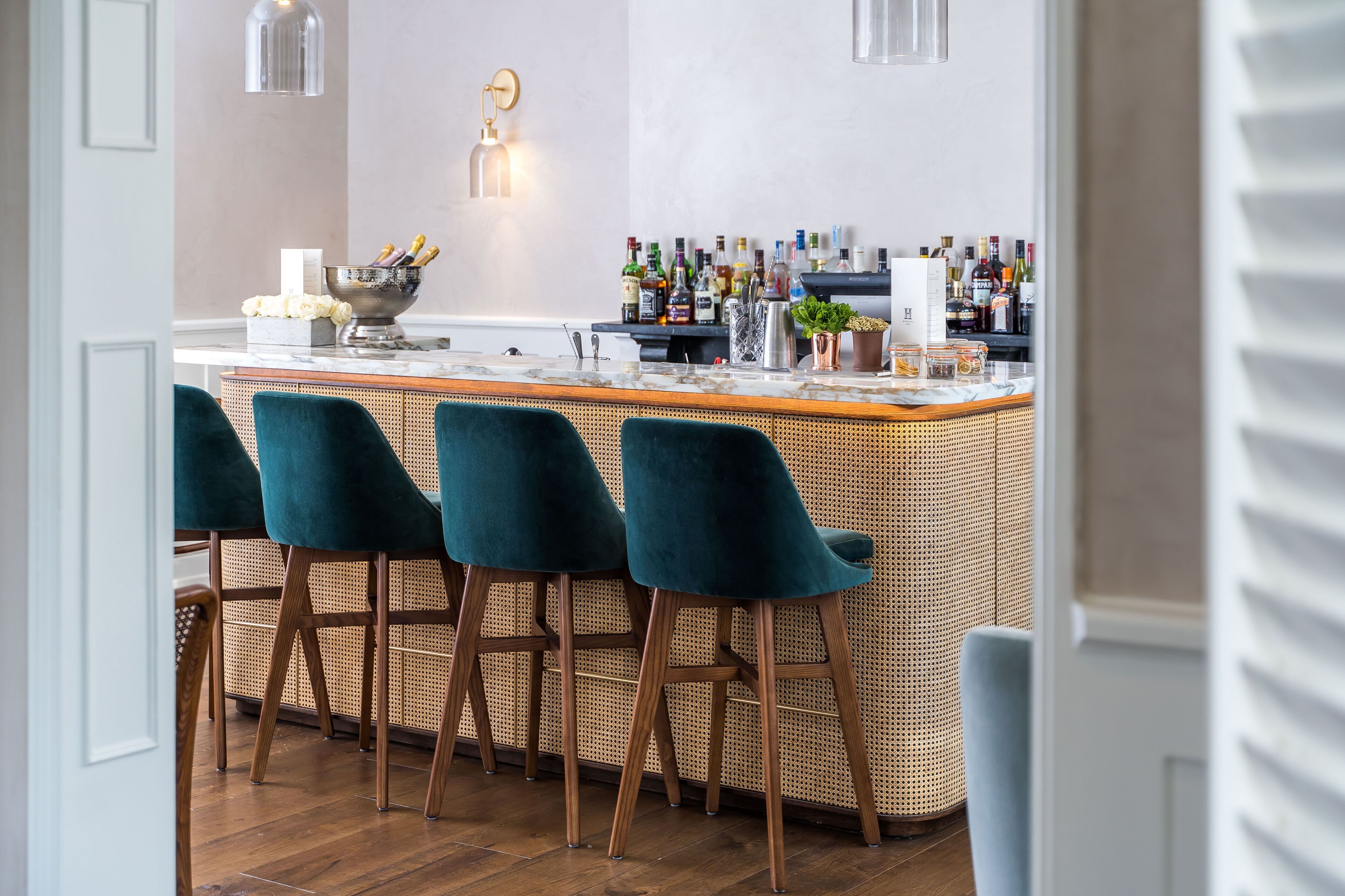 A bar area with a wooden bar counter, green velvet bar stools, and a variety of bottles and glasses.