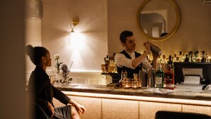 A bar area with a bartender preparing a drink, a seated guest, and a variety of bottles and glasses on the counter.