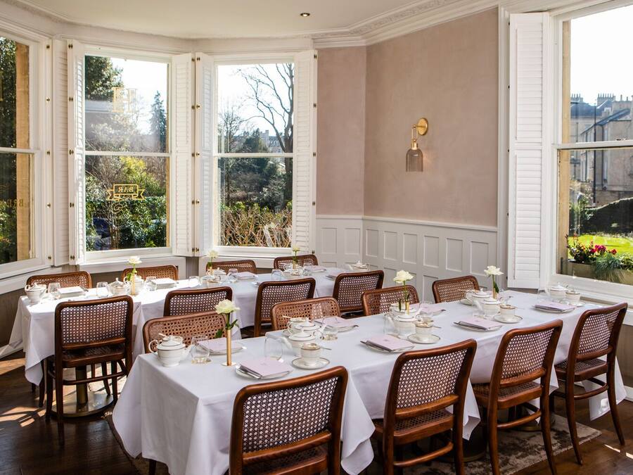 Comedor con una larga mesa dispuesta para una comida, rodeado de sillas de mimbre, con vistas a espacios verdes y edificios a través de amplias ventanas.